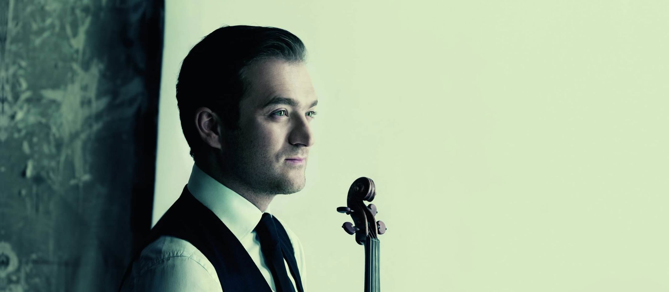 Violinisten Renaud Capuçon i klassisk jakkesæt med slips og sin violin mellem begge hænder foran hvid baggrund.