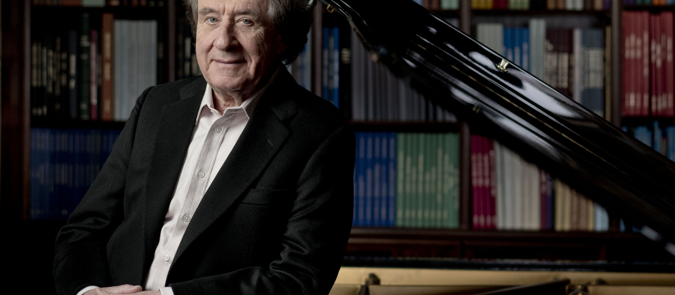 Rudolf Buchbinder læner op ad reflekterende klaver foran stor boghylde iført klassisk jakkesæt.