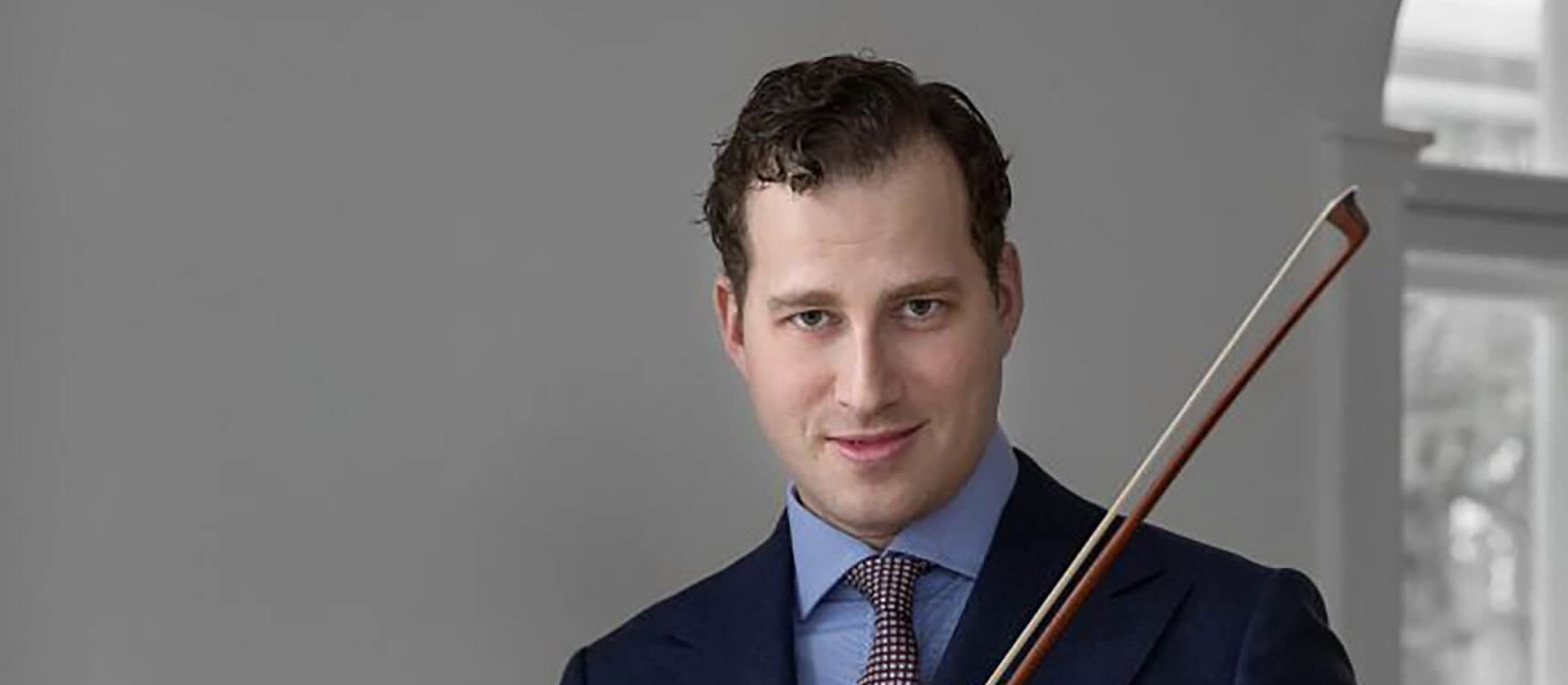 Nikolaj Szeps Znaider med fin violin i hånden foran hvid væg og vindue iført lyseblå skjorte og mørkeblå jakke udover og gå bukser.