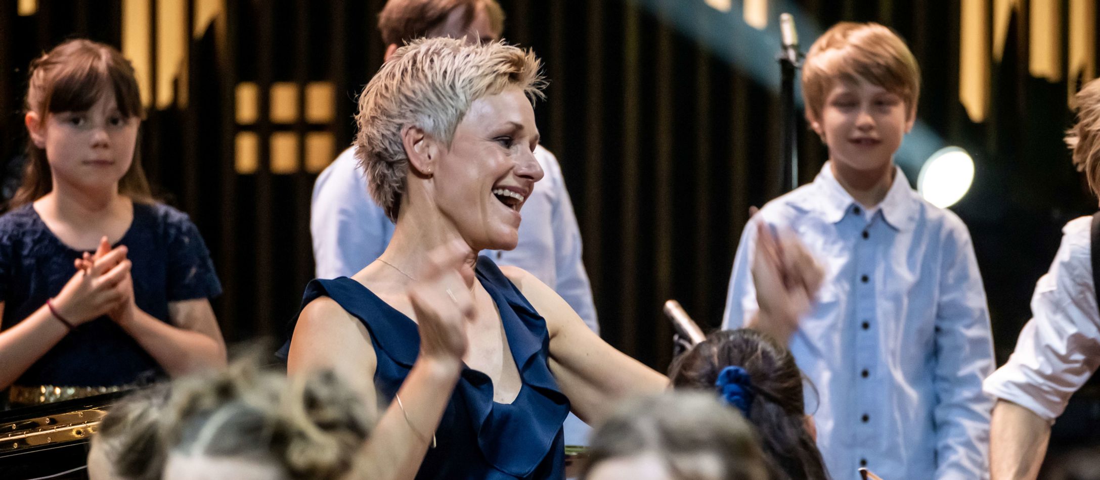 Dirigenten Susanne Wendt smiler og danser i blå kjole mens hun dirigerer DR børnekoret