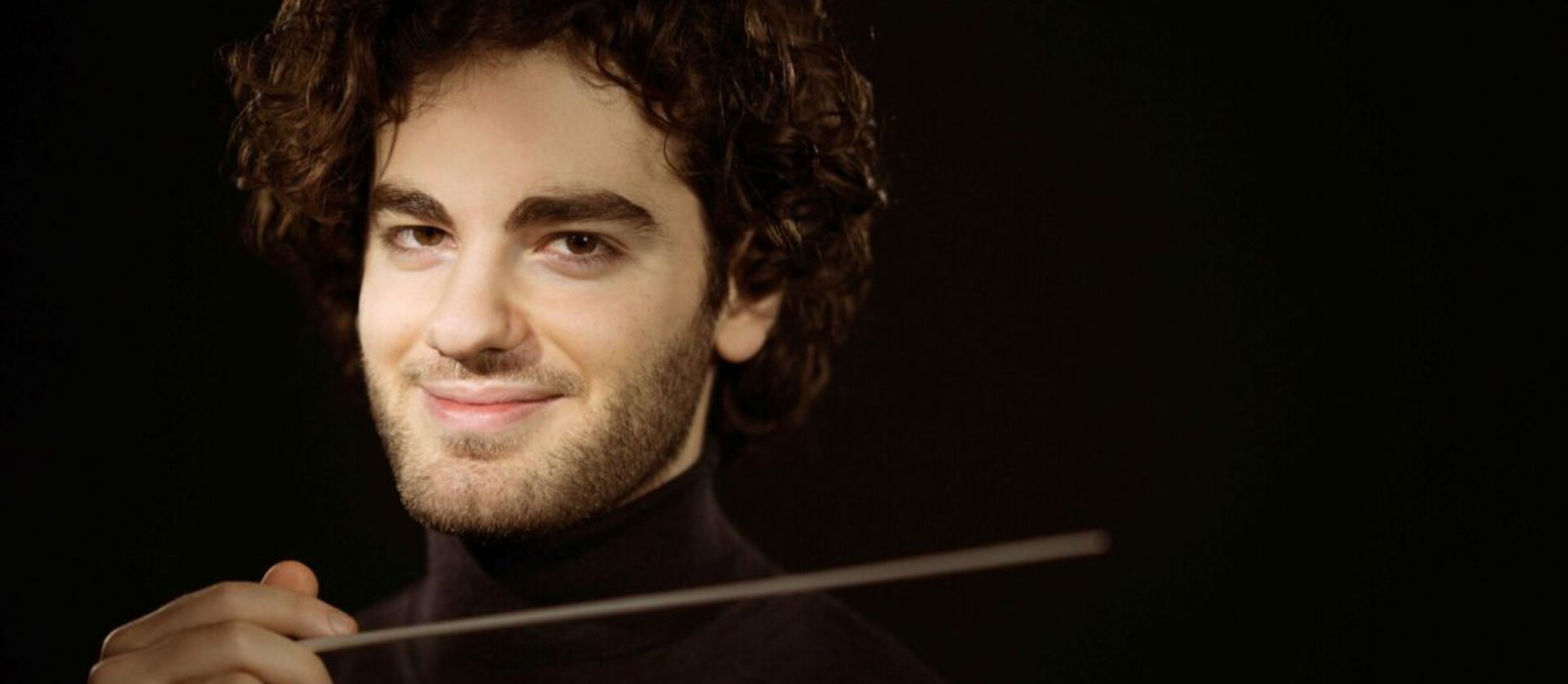 smilende Emmanuel Tjeknavorian med dirigentstang i højrehånd og kruset, krøllet hår og brune øjne.
