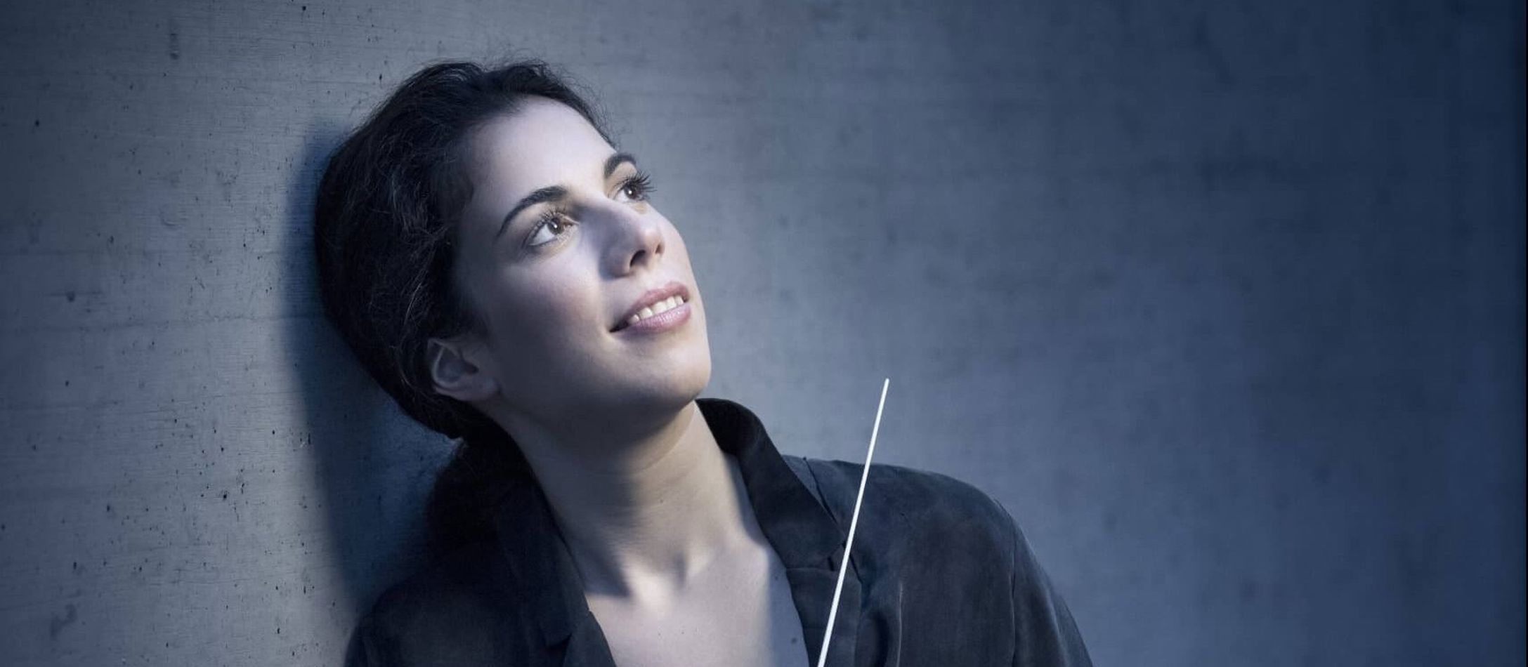 Marie Jacquot med dirigentstang i den ene hånd smiler mens hun læner sig op ad grå mur og kigger opad.