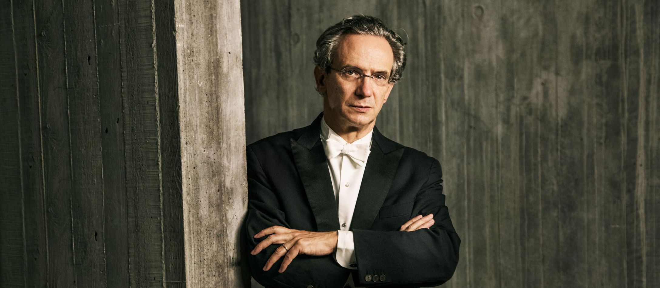 Fabio Luisi med kortlagte arme iført elegant gammeldags habit læner sig opad træværk