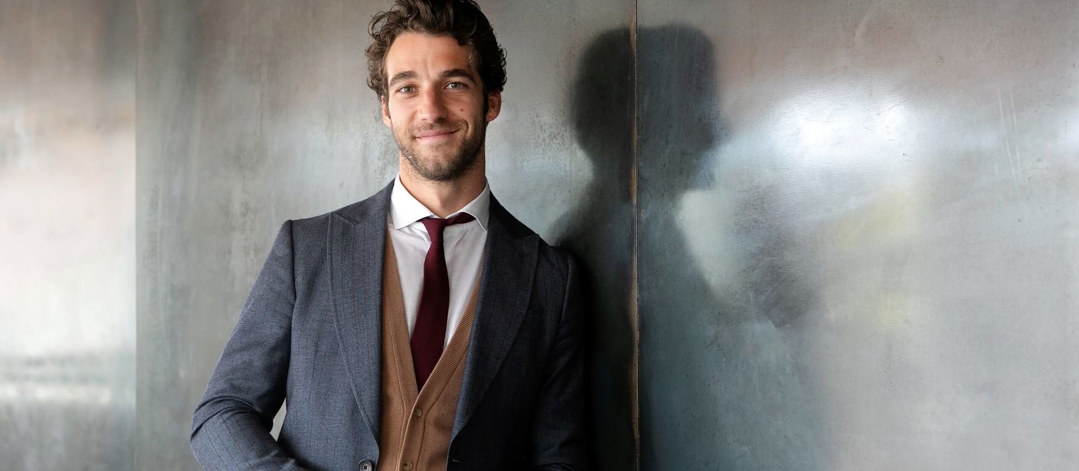 Letsmilende og varme mørkhårede Lorenzo Viotti læner op ad reflekterende væg iført gråt jakkesæt med rødligt slips.