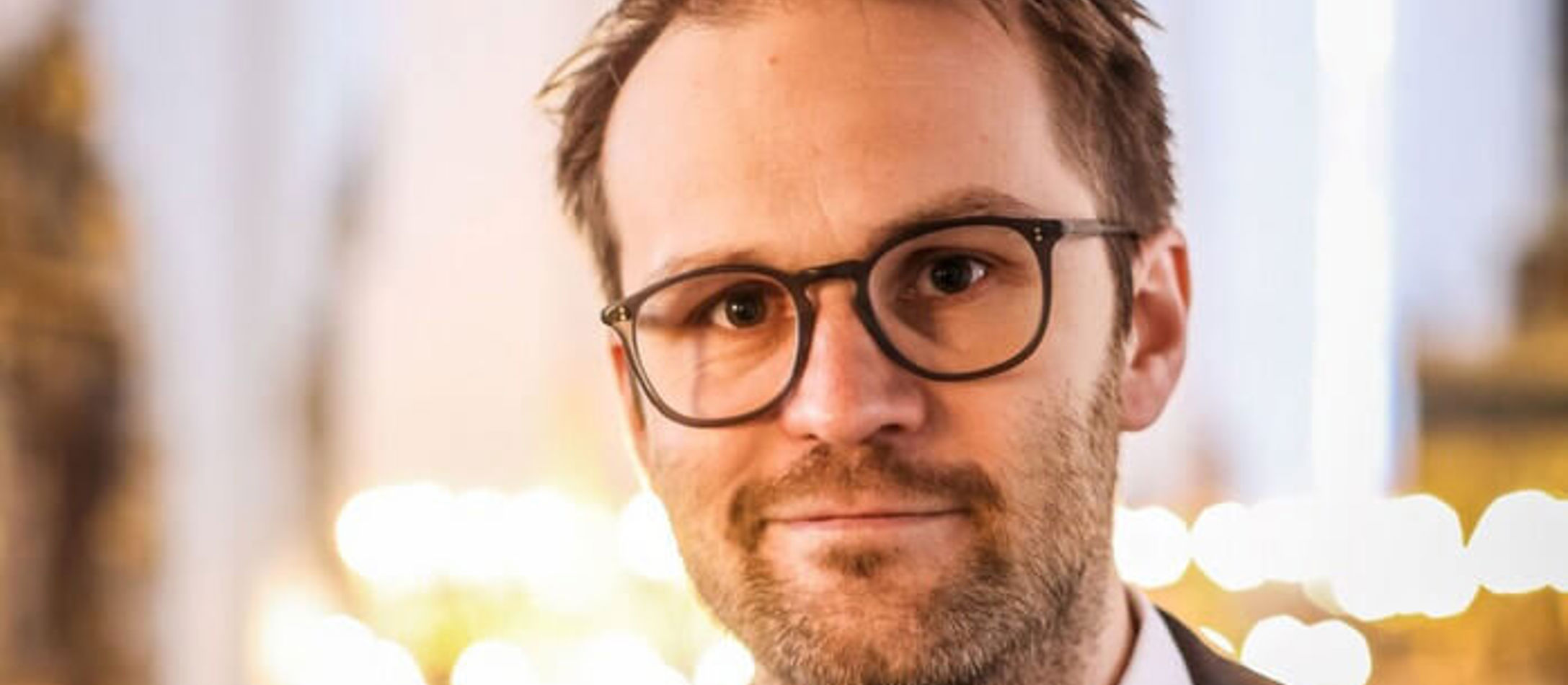 Kristian Krogsøe med sort briller og et skævt lille smil foran lys sløret baggrund.